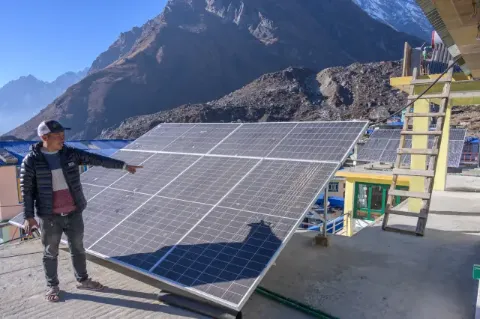Ein Mann steht auf einem betonierten Fläche vor einem Solarpanel und zeigt darauf. Im Hintergrund ist eine alpine Berglandschaft zu sehen.