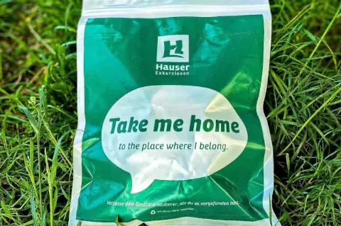 Ein grün bedruckter Beutel liegt auf einer Wiese. Auf dem Beutel ist das Logo von Hauser Exkursionen sowie der Satz "Take me home to the place where I belong" aufgedruckt.