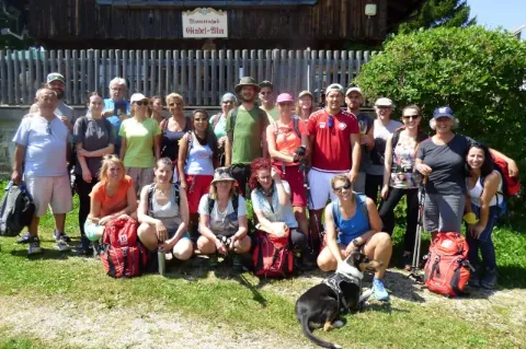 Das Hauser-Team steht und sitzt in Wanderkleidung auf einer grünen Wiese vor einer Hütte und schaut in Richtung Kamera