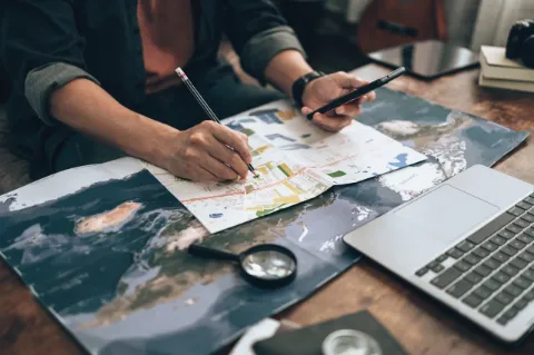 Auf einem Tisch liegen eine Landkarte, ein Laptop und eine Lupe. Eine Person hält einen Bleistift und ein Handy in der Hand.