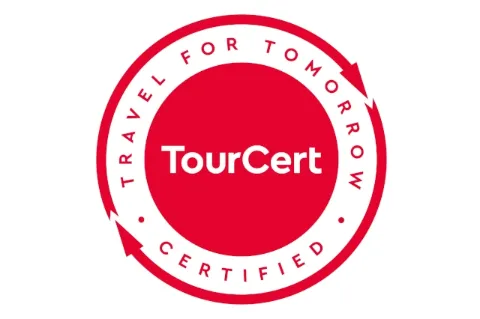Vor weißem Hintergrund ist ein roter Kreis zu sehen in dem in weißer Schrift "TourCert" steht. Um den Kreis stehen die Worte "Travel for Tomorrow certified". Um die Schrift sind kreisförmige Pfeile abgebildet.