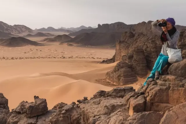 Eine Frau mit Kopftuch sitzt mit einer Kamera rechts vorne im Bild auf einem großen Felsen. Im Hintergrund ist eine Sandwüste zu sehen. Immer wieder ragen große Felsen aus dem Sand hervor. Die Frau blickt durch das Objektiv der Kamera.