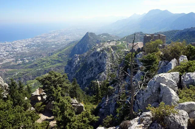 Blick von oben über alte und bewachsene Ruinen, die zwischen bewachsenen Felsen steht. Im Hintergrund ist eine Ebene zu sehen.