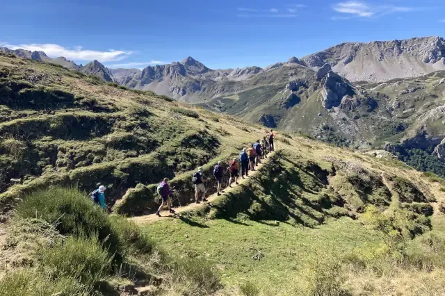 Eine Gruppe Wanderer geht auf einem schmalen Pfad auf einem grünen Hügel. Im Hintergrund sind Felsen zu sehen.