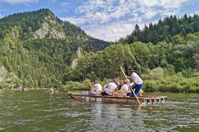 Eine kleine Gruppe sitzt auf einem hölzernen, stabilen Floß. Hinten steht ein Flößer mit einem Stock. Das Floß treibt auf einem grünblauen Fluss. Im Hintergrund ist Nadelwald zu sehen. 