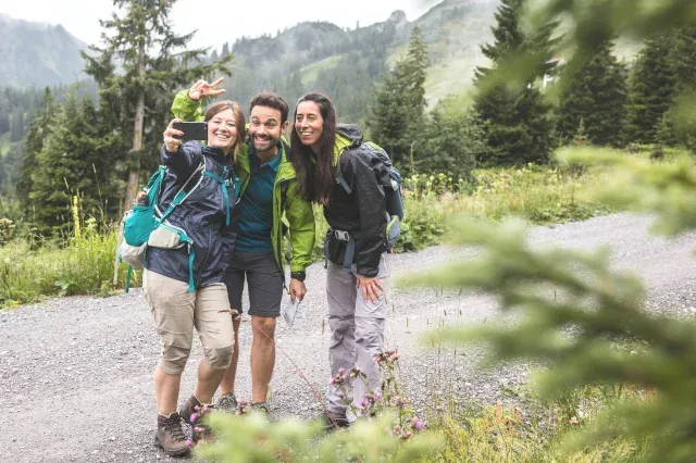 Drei junge Wanderer stehen auf einem Weg und blicken in eine Smartphone-Kamera.