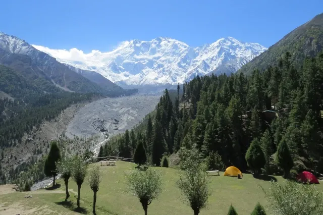 Im Vordergrund ist eine grüne Wiese mit zwei Zelten zu sehen. Hinter einer felsigen Tallandschaft ist das schneebedeckte Bergmassiv des Nanga Parbat zu sehen.
