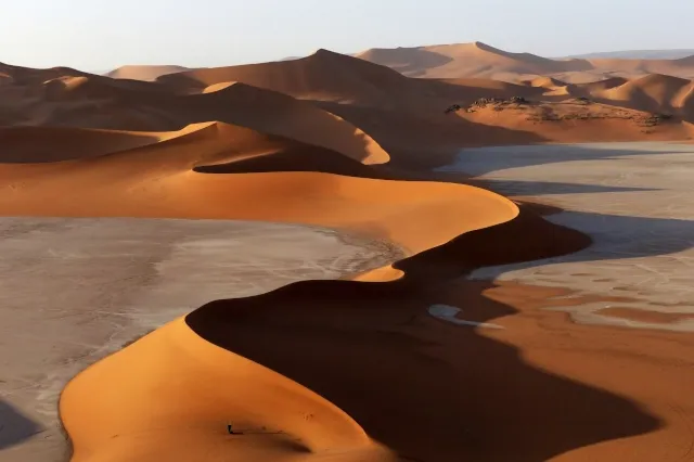 Blick auf eine geschwungene, steil zulaufende Düne, die in Kurven verläuft. Auf der rechten Seite der Düne wirft diese einen großen Schatten. Im Hintergrund sind weitere Sanddünen zu sehen.