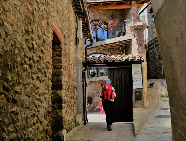 Eine Frau mit rotem Rucksack geht durch ein Tor in einer engen Gasse mit Steinhäusern.
