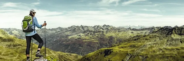 Eine Frau mit Rucksack und Wanderstöcken schaut auf eine Gebirgslandschaft hinunter.