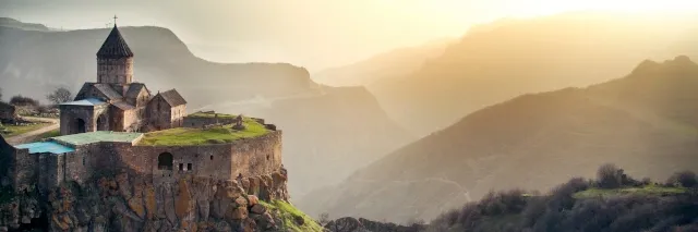 Blick auf das Tatev Kloster das direkt auf einen hohen Felsen gebaut ist und vor einer Berglandschaft liegt. Im Hintergrund geht die Sonne unter.