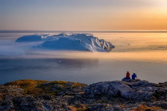 Zwei Menschen sitzen auf einem Felsen und schauen auf das Meer hinaus. Auf dem Meer ist der Teil eines Gletschers zu sehen und am Horizont geht die Sonne unter.