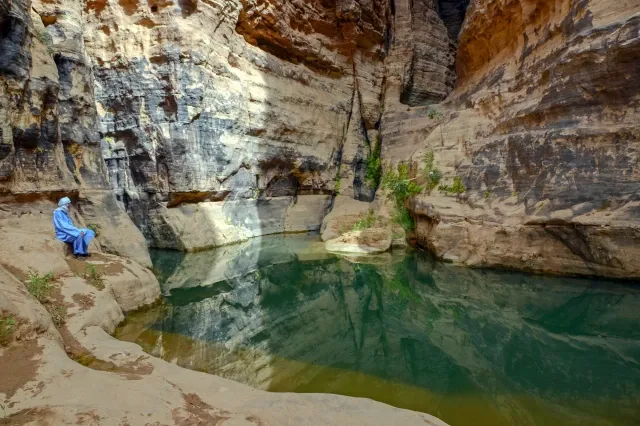Ein Tuareg sitzt an einem kristallklarem Bergsee und schaut auf den See. Die Bergwände sind farbig.