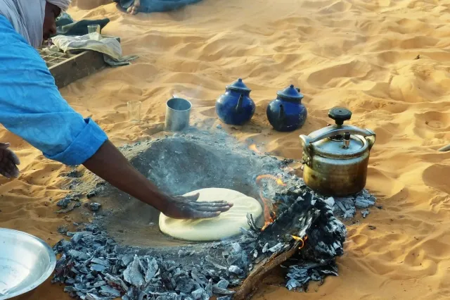 Ein Tuareg bereitet Fladenbrot mit seiner Hand in einer heißen Grube im Sand, gefüllt mit Holzkohle, zu.