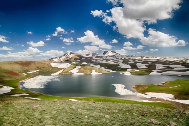 Ein See in der Mitte. Darum sind Berge und ein grün-orangenes Tal. Stellenweise gibt es Schneefelder.