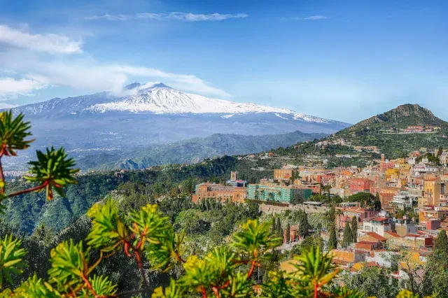 Die Stadt Taormina am Hang eines Hügels. Im Hintergrund befindet sich der schneebedeckte Ätna.