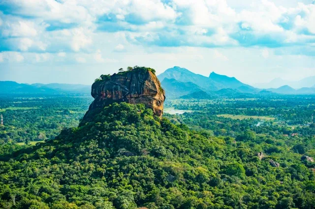 Eine weite Waldlandschaft, im Hintergrund sind Berge. In der Mitte ist ein großer eckiger Felsen.