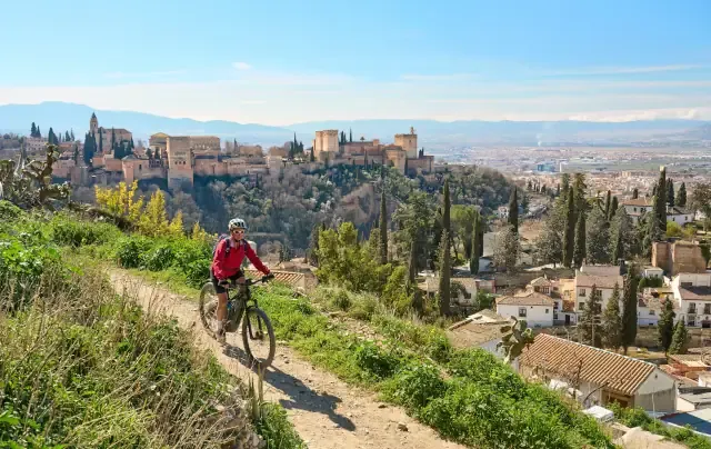 Eine Frau radelt mit ihrem E-Bike auf einer Schotterpiste. Im Hintergrund sieht man eine Stadt und eine Burg.