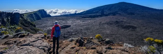 Wanderer steht am Kraterrand eines Vulkans und blickt auf die schwarze Lavalandschaft im Krater