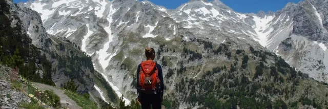 Eine Wanderin mit rotem Rucksack steht vor einem gräulichen, teils schneebdeckten Bergmassiv.