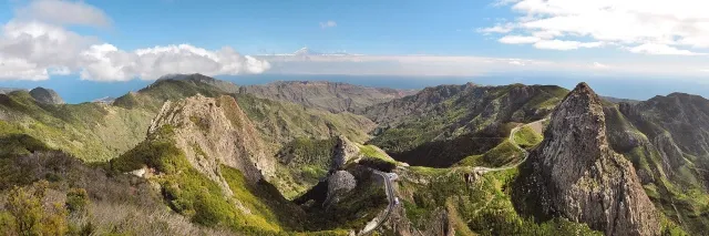 Ein zerklüftetes Bergpanorama. Die Berge sind grün bewachsen. In der Ferne ist das Meer.