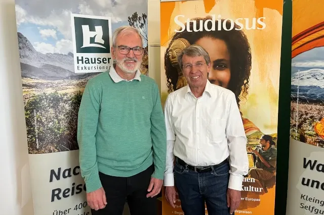 Zwei Männer stehen vor zwei Werbeflächen der Firmen Studiosus und Hauser Exkursionen. Beide schauen in die Kamera und lächeln.