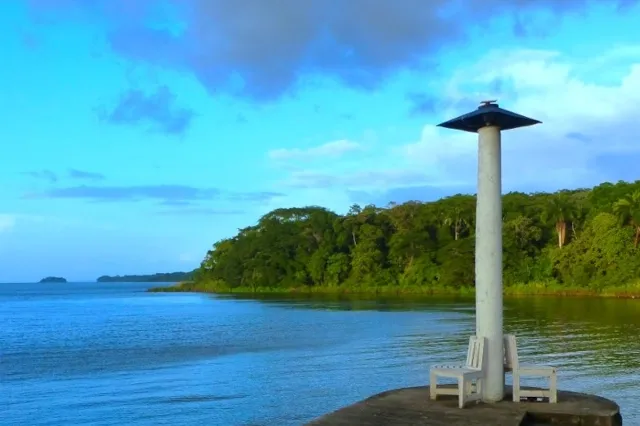 Ein kleiner grauer Leuchtturm steht auf einem Plateau auf dem Wasser. Im Hintergrund ist ein Teil einer grünen Insel zu sehen.