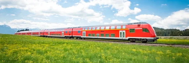 Eine rote Regionalbahn fährt auf Gleisen in einer blumigen Naturlandschaft.