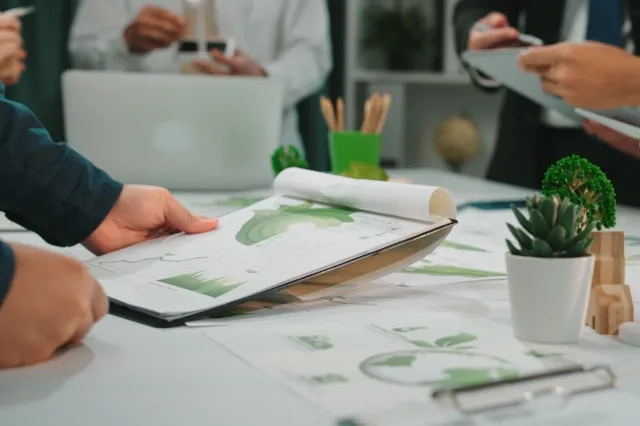 Szene einer Geschäftsbesprechung mit Fokus auf Nachhaltigkeit. Auf dem Tisch liegen Dokumente mit verschiedenen Diagrammen. Auf dem Tisch steht eine kleine Pflanze sowie weitere Büromaterialien.