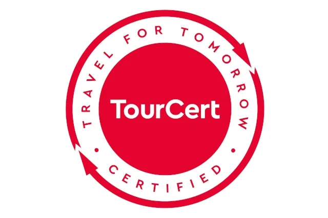 Vor weißem Hintergrund ist ein roter Kreis zu sehen in dem in weißer Schrift "TourCert" steht. Um den Kreis stehen die Worte "Travel for Tomorrow certified". Um die Schrift sind kreisförmige Pfeile abgebildet.