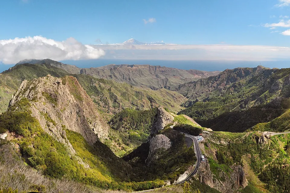 Ein zerklüftetes Bergpanorama. Die Berge sind grün bewachsen. In der Ferne ist das Meer.