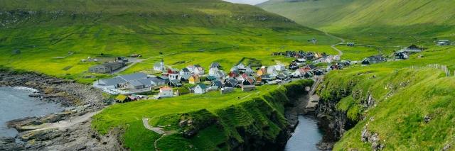 Luftaufnahme von Dorf an der Küste umringt von grünen Wiesen und grünen Berghängen
