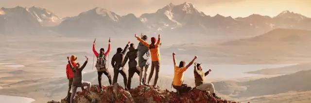 Gruppe von neun Wanderern steht auf dem Gipfel und jubelt in einer Sonnenuntergangsstimmung