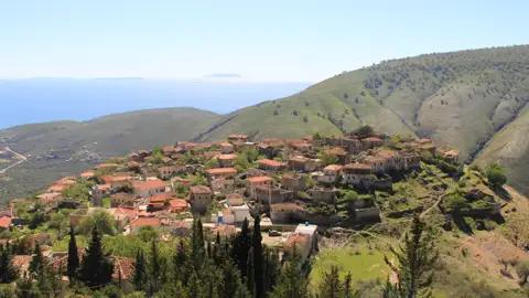 Panoramablick auf ein Bergdorf mit traditionellen Häusern, das sich an einen Hügel schmiegt. Im Hintergrund erstreckt sich das blaue Meer.