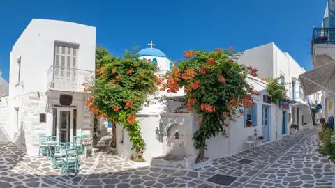 Traditionelle Architektur einer griechischen Inselgasse mit weiß getünchten Häusern, blühenden Pflanzen und einer blau-weißen Kirche.