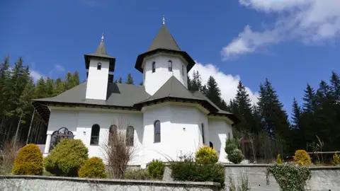 Weiße Kirche mit dunklen Türmen und Dach, umgeben von Nadelbäumen.