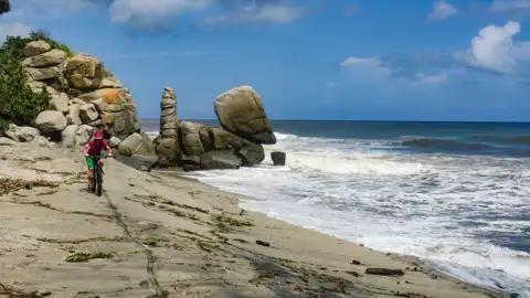 Radfahrer am Strand mit Felsformationen im Hintergrund.