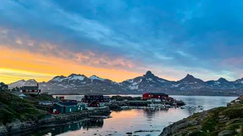 Sonnenuntergang über einem Küstenort und schneebedeckten Bergen in Grönland.