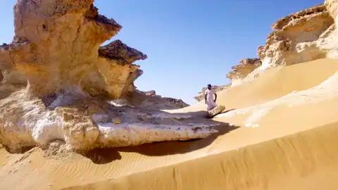Person in traditioneller Kleidung inmitten von Wüstenfelsformationen und Sanddünen.