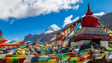 Gebetsfahnen flattern im Wind vor einer Stupa mit dem Berg Kailash im Hintergrund.
