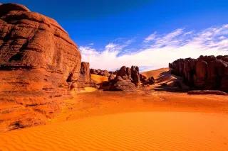 Rötliche Felsen ragen aus dem rötlichen Sand der Wüste hervor. Der Sand ist wellig gezeichnet vom Wind.