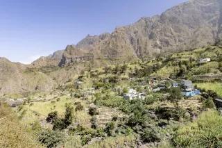Ein kleines Bergdorf am Hang. Die Landschaft ist grün. Im Hintergrund sind bräunliche Berge.