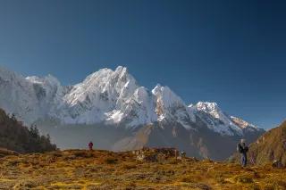 Zwei Wanderer blicken von einem orange-grün bewachsenen Berg auf einen schneebedeckten Berg.