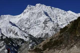 Eine große schneebedeckte Bergwand. Davor ein felsiger Berg. Der Himmel ist blau.