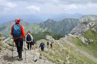 Sechs Personen mit Wanderstöcken laufen hintereinander über einen Grat bei bewölktem Himmel