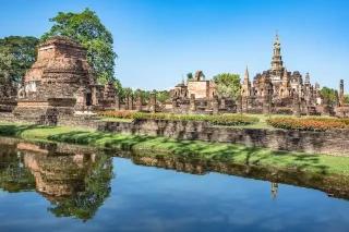 Blick vom Wasser auf einen historischen Park mit kleinen Gebäuden. Um den Park herum wächst viel Grünes.