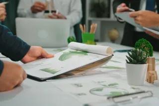 Szene einer Geschäftsbesprechung mit Fokus auf Nachhaltigkeit. Auf dem Tisch liegen Dokumente mit verschiedenen Diagrammen. Auf dem Tisch steht eine kleine Pflanze sowie weitere Büromaterialien.