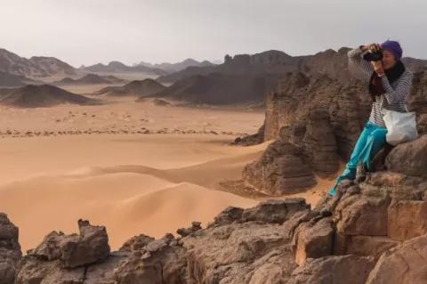 Eine Frau mit Kopftuch sitzt mit einer Kamera rechts vorne im Bild auf einem großen Felsen. Im Hintergrund ist eine Sandwüste zu sehen. Immer wieder ragen große Felsen aus dem Sand hervor. Die Frau blickt durch das Objektiv der Kamera.