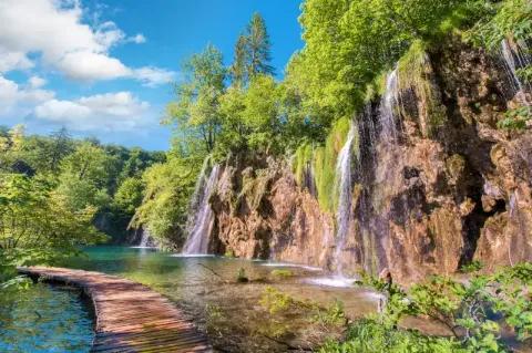 Holzsteg führt durch Wasser vorbei an Wasserfällen mit grünen Bäumen oberhalb