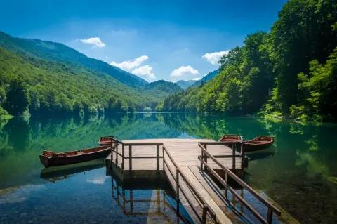 Ein hölzerner Steg mit Plattform vorne ragt in einen dunkelblauen See. Den See umrahmen bewaldete Hügel.